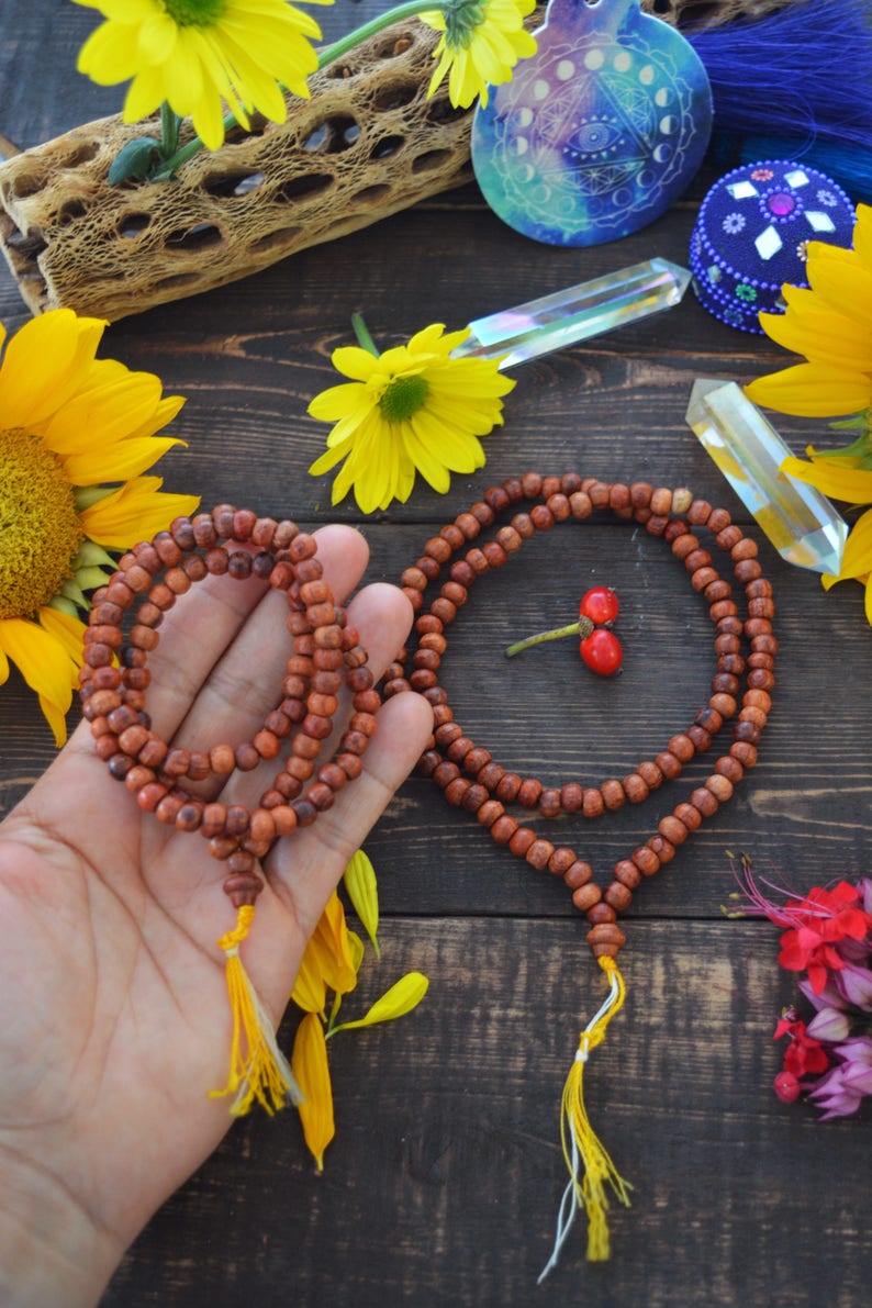May include: Two brown wooden mala bead necklaces with yellow tassels. The necklaces are on a wooden surface with yellow flowers and other decorative items.