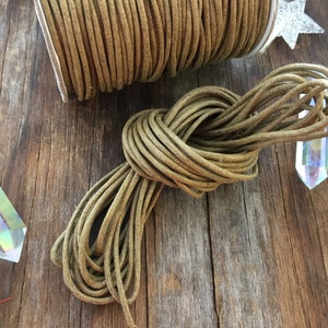 May include: A spool of olive green cord sits next to a knotted bundle of the same cord. The cord is made of a round, braided material. The background includes a wooden surface and two iridescent crystal prisms.