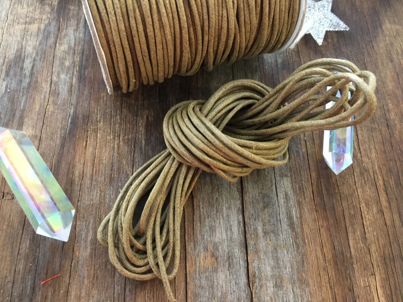 May include: A spool of olive green cord sits next to a knotted bundle of the same cord. The cord is made of a round, braided material. The background includes a wooden surface and two iridescent crystal prisms.