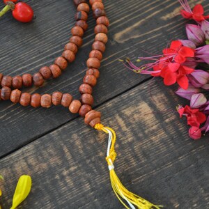 May include: A wooden mala bead necklace with a yellow tassel. The necklace is made of brown beads and is lying on a dark wooden surface. There are pink and red flowers in the background.