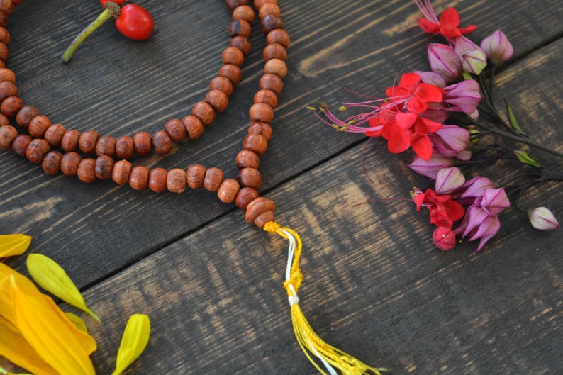May include: A wooden mala bead necklace with a yellow tassel. The necklace is made of brown beads and is lying on a dark wooden surface. There are pink and red flowers in the background.