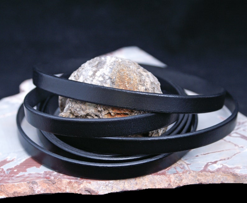 Puede incluir: Cord&oacute;n de cuero negro, enrollado sobre una superficie de piedra. El cord&oacute;n tiene aproximadamente 6 mm de ancho.