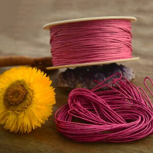 May include: A spool of pink waxed cotton cord, with a loose end of the cord in the foreground. The cord is wound around a white cardboard spool.
