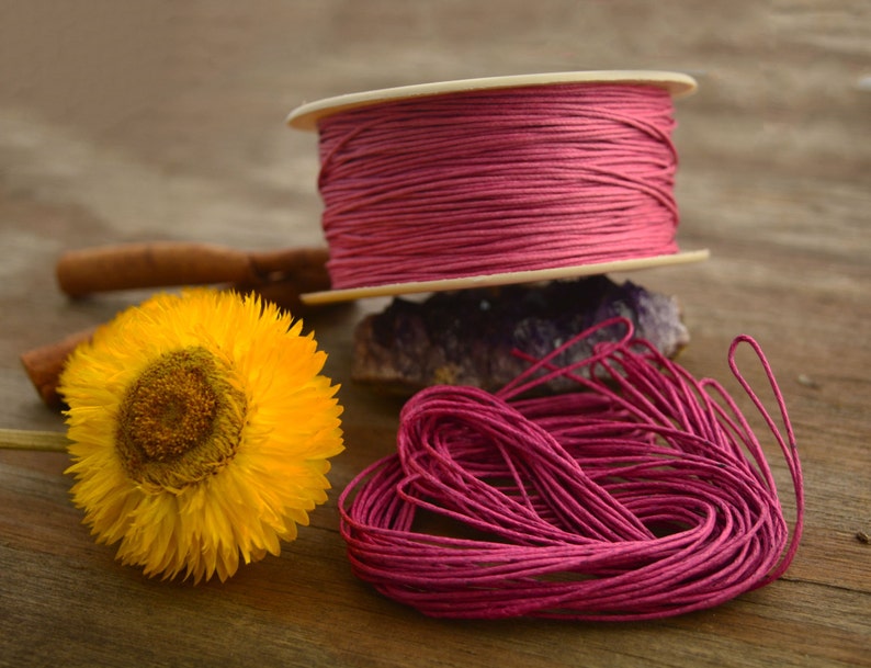 May include: A spool of pink waxed cotton cord, with a loose end of the cord in the foreground. The cord is wound around a white cardboard spool.