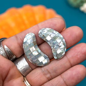 May include: A pair of crescent-shaped earrings made of iridescent mother-of-pearl tiles. The tiles are arranged in a checkered pattern.
