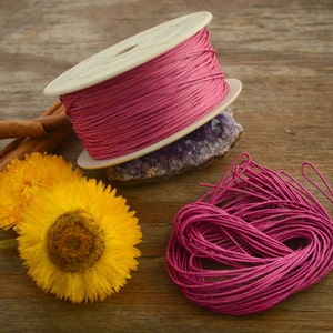 May include: A spool of pink waxed cotton cord, wound around a white plastic spool, with a loose end of the cord hanging down. The cord is also shown in a loose pile next to the spool.