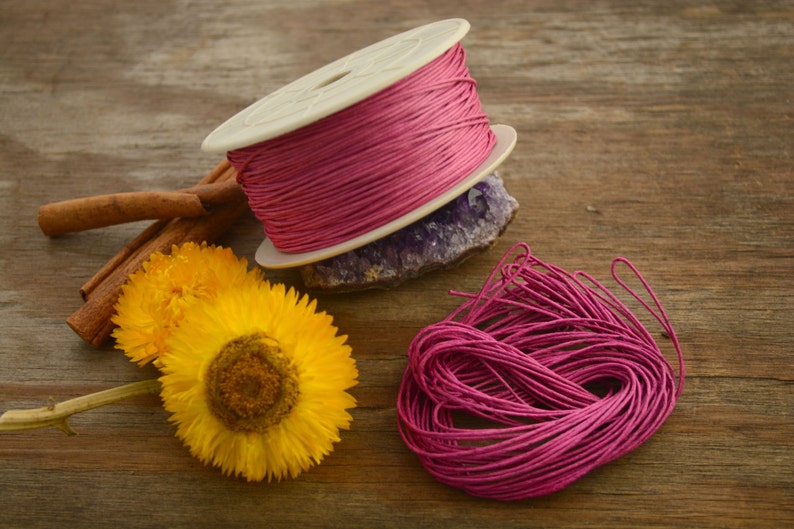 May include: A spool of pink waxed cotton cord, wound around a white plastic spool, with a loose end of the cord hanging down. The cord is also shown in a loose pile next to the spool.