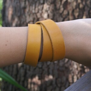 Puede incluir: Pulsera envolvente de cuero amarillo mostaza. La pulsera está enrollada alrededor de la muñeca, mostrando la textura suave y el color cálido del cuero. Un accesorio sencillo pero elegante.