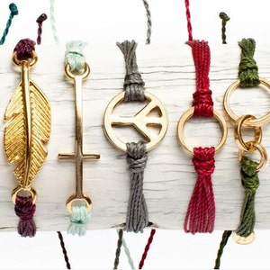 Peut inclure: Un ensemble de huit bracelets en corde de couleur avec des breloques dor&eacute;es. Les breloques comprennent une plume, une croix, un signe de paix, un cercle, un symbole d'infini et deux autres cercles.