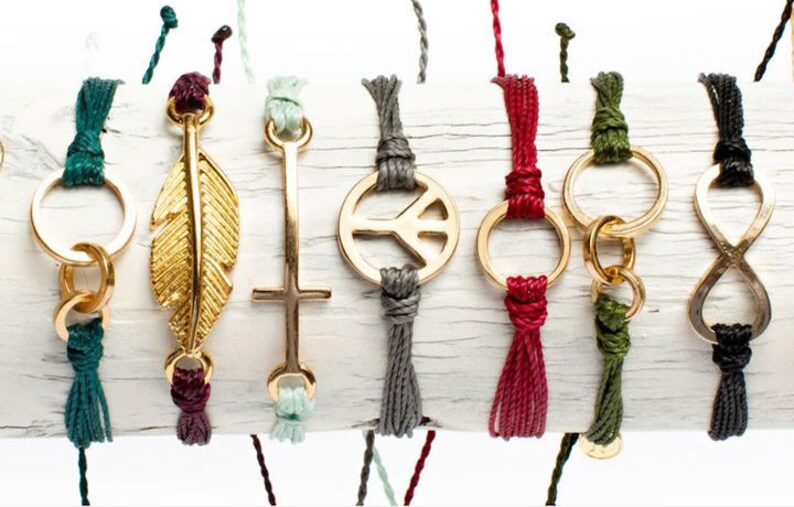 Peut inclure: Un ensemble de huit bracelets en corde de couleur avec des breloques dor&eacute;es. Les breloques comprennent une plume, une croix, un signe de paix, un cercle, un symbole d'infini et deux autres cercles.