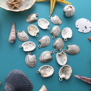 May include: A collection of silver-toned shell-shaped charms, perfect for jewelry making or crafting. The charms are arranged on a blue background with seashells and starfish.