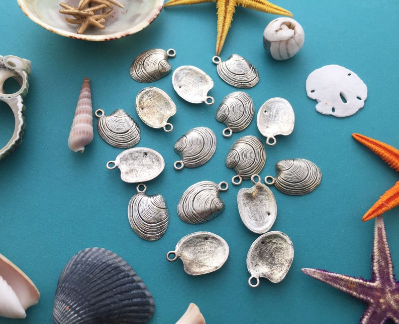 May include: A collection of silver-toned shell-shaped charms, perfect for jewelry making or crafting. The charms are arranged on a blue background with seashells and starfish.
