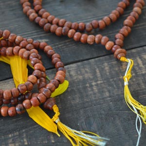 May include: Two brown wooden beaded necklaces with yellow tassels. The necklaces are on a wooden surface with yellow flower petals.