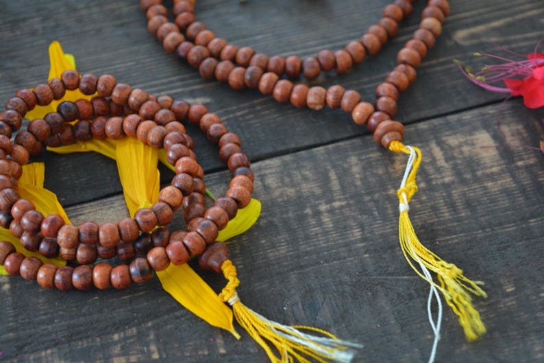 May include: Two brown wooden beaded necklaces with yellow tassels. The necklaces are on a wooden surface with yellow flower petals.