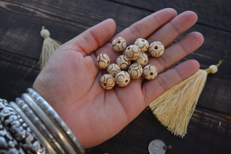May include: A hand holding a collection of white wooden beads with brown circular patterns. The beads are approximately 1 inch in diameter.