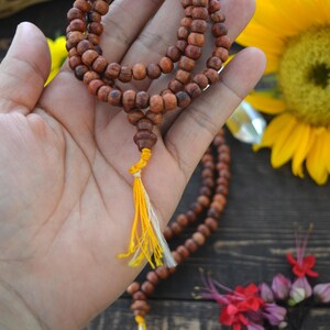 May include: A brown wooden mala bead necklace with a yellow and white tassel. The necklace is held in a hand against a wooden background with sunflowers and other flowers.
