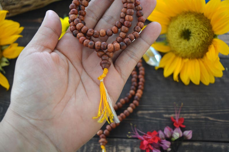 May include: A brown wooden mala bead necklace with a yellow and white tassel. The necklace is held in a hand against a wooden background with sunflowers and other flowers.
