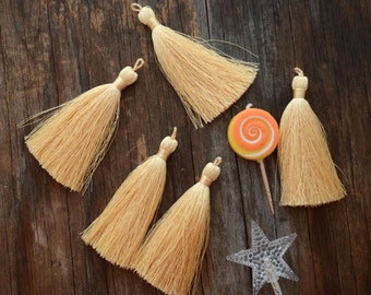 May include: Five light beige tassels with looped tops are arranged on a dark wood surface. A lollipop with orange and yellow swirls and a clear star-shaped light are also present. The tassels are made of a soft, flowing material.