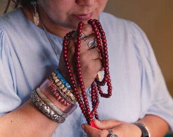 May include: A person wearing a wide-brimmed hat and a light blue shirt holds a long strand of red wooden beads. The beads are wrapped around their hand and wrist, and they are wearing several bracelets on their wrist.