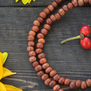 May include: A wooden mala necklace with brown beads on a dark wood background. Yellow flowers and red berries are also in the image.