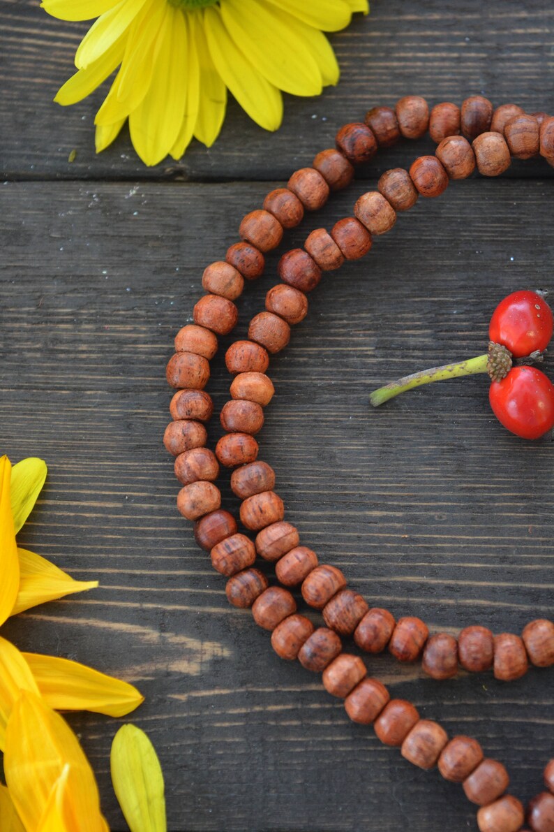May include: A wooden mala necklace with brown beads on a dark wood background. Yellow flowers and red berries are also in the image.