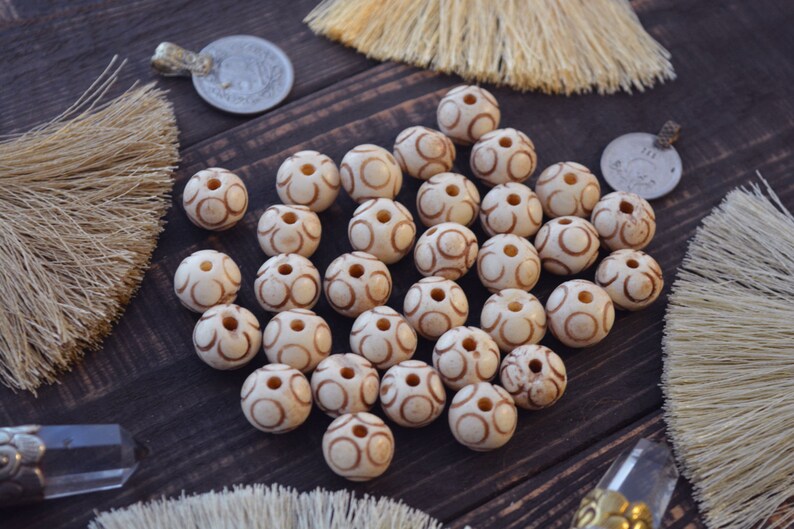 May include: A collection of white beads with brown swirl designs. The beads are arranged on a dark brown wooden surface.