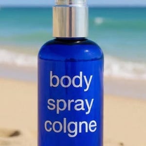 Puede incluir: Una botella de colonia en spray para el cuerpo de vidrio azul con detalles plateados y una tapa transparente. Las palabras "body spray cologne" están impresas en blanco. La botella está sobre una roca, con una playa y el mar de fondo.