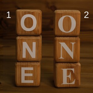 May include: Two stacks of wooden blocks spelling the word "ONE". Each block is a natural wood color with white letters. The blocks are arranged on a wooden surface.