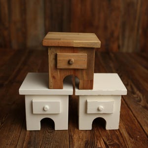 May include: Three small wooden stools. One is natural wood with a drawer and a round knob. The other two are painted white with a drawer and a round knob. The stools have a curved cutout at the base.