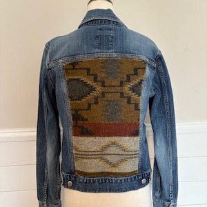 May include: Blue denim jacket with a patterned fabric panel on the back. The panel features geometric designs in shades of brown, gray, and red. The jacket has a classic collar, button closures, and pockets.