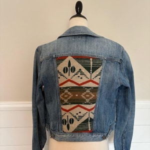 May include: A light blue denim jacket with a decorative patterned panel on the back. The panel features a geometric design in shades of red, green, brown, and cream. The jacket has a classic collar and button closures.