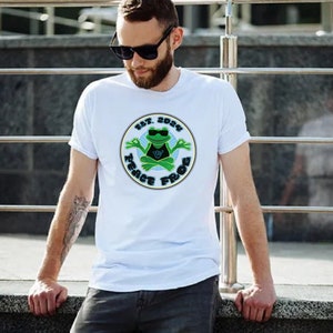 Puede incluir: Camiseta blanca con una rana verde que lleva gafas de sol y medita en un círculo con el texto "Est. 2024 Peace Frog"
