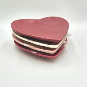 Puede incluir: Una pila de platos de cerámica en forma de corazón en tonos rosa y blanco. Los platos están apilados uno encima del otro, con un plato de color rosa intenso en la parte superior y un plato rosa claro en la parte inferior. Sobre un fondo blanco.