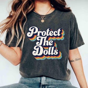 Puede incluir: Una camiseta gris oscuro con un gráfico de estilo vintage que dice "Protect The Dolls" en colores arcoíris.
