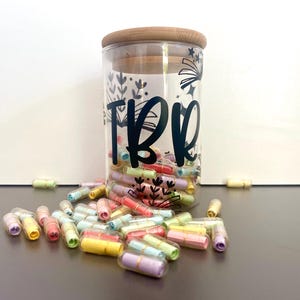 Op de afbeelding: Een glazen pot met een bamboe deksel gevuld met kleurrijke capsules. De pot heeft een zwart-wit ontwerp met het woord "TRB" erop.