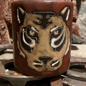 May include: A cylindrical, brown ceramic mug featuring a detailed tiger face design. The tiger's face is tan with black and white accents, set against the dark brown background of the mug. The mug has a rustic, handcrafted appearance.