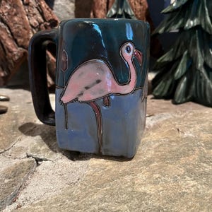 Op de afbeelding: Een keramische mok met een flamingo-ontwerp. De mok heeft een donkerblauwe achtergrond met een roze flamingo-illustratie. De flamingo heeft een rode snavel en staat in blauw water. De handgreep is zwart.