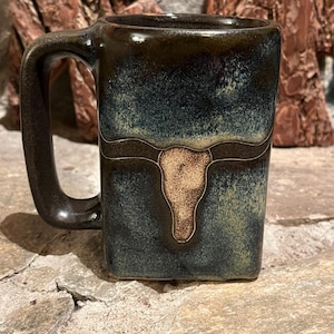 May include: A square-shaped ceramic mug with a dark blue and brown glaze. The mug features a tan bull skull design. The handle is a dark brown color. The mug is sitting on a stone surface.