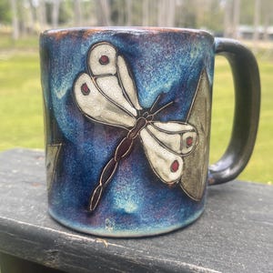 May include: A ceramic mug with a dragonfly design. The mug has a blue and brown glaze with a raised dragonfly in cream and brown. The dragonfly has red accents on its wings. The handle is dark brown.