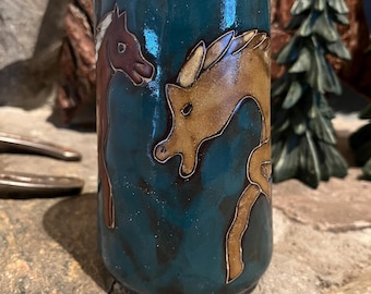 May include: A teal ceramic tumbler with brown and tan horse and deer designs. The tumbler has a cylindrical shape and is decorated with a glossy finish. The animals are outlined in a darker brown, creating a contrasting effect against the teal background.