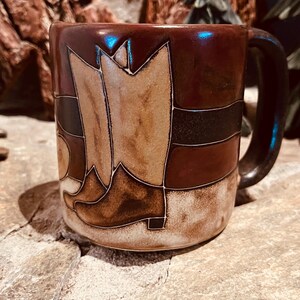 Op de afbeelding: Een keramische mok met een ontwerp van bruine en beige cowboylaarzen. De mok heeft een donkerbruin handvat en een glanzende afwerking. De laarzen zijn beige met bruine accenten, tegen een achtergrond van bruine en beige strepen.