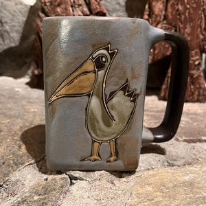 May include: A ceramic mug with a square shape and a dark brown handle. The mug is a muted blue-gray color and features a detailed illustration of a pelican in shades of brown and green. The mug is sitting on a stone surface.