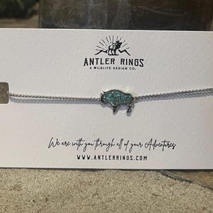 Op de afbeelding: Een zilverkleurige kettingarmband met een centrale bedel in de vorm van een bizon met een getextureerde, blauwgroene inleg. De armband wordt gepresenteerd op een witte kaart met de tekst Antler Rings, A Wildlife Design Co., de zin We are with you through all of your adventures en de website-URL www.antlerrings.com.