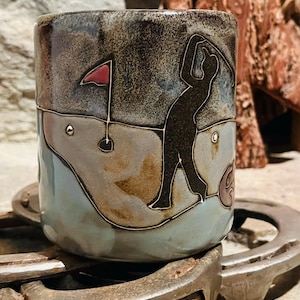 Peut inclure: Mug en céramique sur le thème du golf avec un glaçage marron foncé et bleu. La tasse présente une silhouette de golfeur, un drapeau rouge et une balle de golf. La tasse est posée sur un fer à cheval.