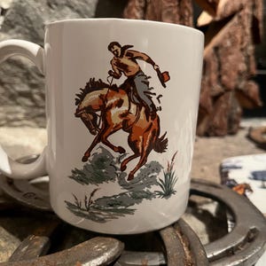 Op de afbeelding: Witte keramische mok met een vintage illustratie van een cowboy te paard. De cowboy draagt een bruin overhemd en een grijze broek. Het paard is bruin en wit. De mok heeft een groot handvat en is omringd door hoefijzers.