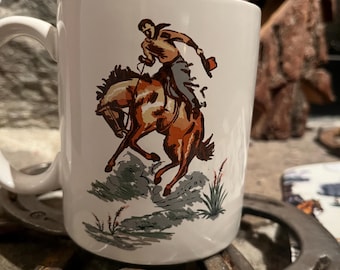 May include: White ceramic mug featuring a vintage illustration of a cowboy riding a horse. The cowboy is wearing a brown shirt and gray pants. The horse is brown and white. The mug has a large handle and is surrounded by horseshoes.