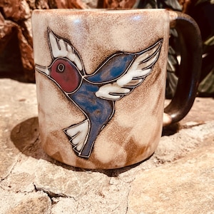 May include: A ceramic mug with a brown handle and a hummingbird design. The hummingbird is depicted with blue, red, and white details against a beige background. The mug is sitting on a stone surface.