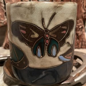 May include: A ceramic mug with a butterfly design. The mug features a brown butterfly with teal, pink, and blue accents against a light beige background. The bottom of the mug is dark blue.