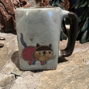 May include: A ceramic mug with a light blue glaze and a brown handle. The mug features a cartoon cat design with a pink body, brown head, and blue tail. The mug is sitting on a stone surface.