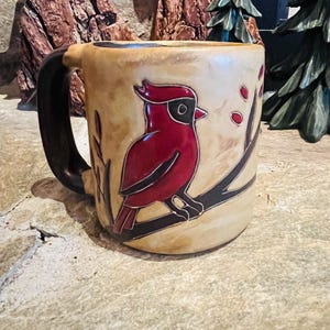 May include: A ceramic mug with a hand-painted cardinal bird design. The mug features a tan background with a dark brown handle and a red cardinal perched on a branch. The mug is approximately 4 inches tall.